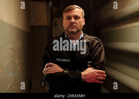 Seriöser, reifer männlicher Wachmann mit einem Haufen Schlüssel in der Hand, Arme an der Brust überkreuzen und in die Kamera blicken, während er im Flur steht Stockfoto