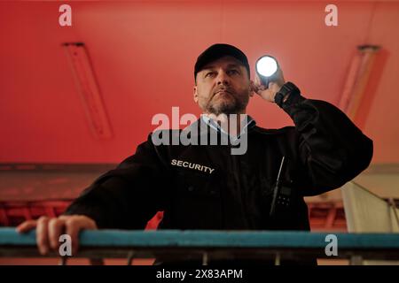 Unter dem Blick auf einen seriösen männlichen Sicherheitsbeamten in einheitlicher Beleuchtung mit Taschenlampe, während er am Geländer steht und den Raum beobachtet Stockfoto