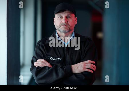 Reifer Wachmann in schwarzer Uniform, der vor der Kamera im Flur steht und seine Arme während der Nachtarbeit von der Brust kreuzt Stockfoto