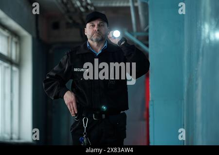 Reifer männlicher Sicherheitsbeamter in Uniform und Beleuchtung nach vorne mit Taschenlampe, während er sich entlang des Korridors des alten Gebäudes bewegt Stockfoto