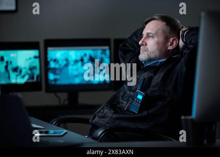 Erholsamer Bediener in Sicherheitsuniform, der die Hände auf dem Hinterkopf hält, während er im Sessel vor dem Computer sitzt und auf den Bildschirm blickt Stockfoto