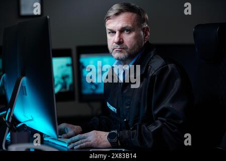 Seriöser und selbstbewusster professioneller Sicherheitsbeamter in Uniform, der die Hände auf Computertastatur und -Maus hält und die Kamera betrachtet Stockfoto