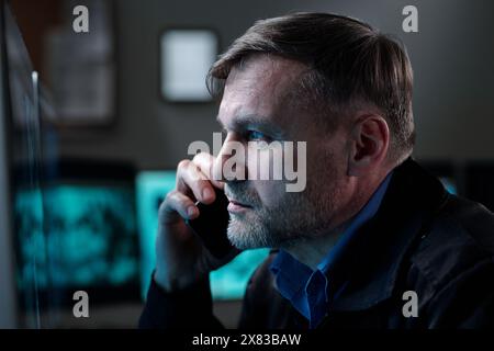 Ernster reifer Mann mit Smartphone am Ohr, der vor dem Computerbildschirm sitzt und cctv-Videos ansieht, während er die Situation dem Kollegen meldet Stockfoto