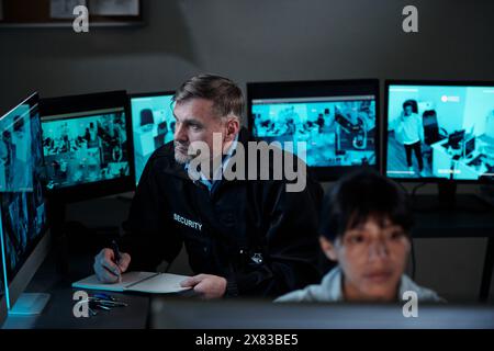 Seriöser, reifer männlicher Wachmann mit Stift über offenem Copybook, der Notizen macht, während er vor dem Computerbildschirm sitzt und cctv-Videos ansieht Stockfoto