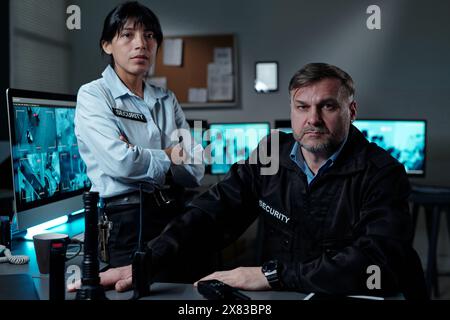 Zwei professionelle interkulturelle Sicherheitsbeamte in Uniform, die in die Kamera blicken, während sie im Überwachungsraum mit Computermonitoren arbeiten Stockfoto