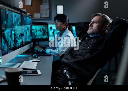 Reifer männlicher Offizier in schwarzer Uniform hält Hände hinter dem Kopf, während er im Sessel vor dem Computerbildschirm mit videoüberwachung sitzt Stockfoto