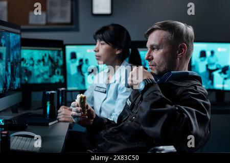 Ein seriöser reifer Offizier in Uniform, der eine Tasse Kaffee und Sandwich trinkt, während er vor dem Computerbildschirm sitzt und cctv-Videos ansieht Stockfoto