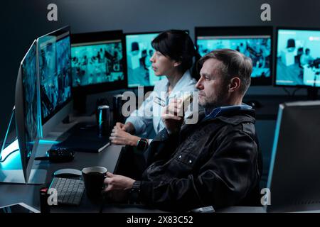 Seitenansicht eines seriösen, Reifen männlichen Sicherheitsbeamten mit Sandwich und einer Tasse Kaffee in den Händen, die videoüberwachung auf dem Computerbildschirm anschauen Stockfoto