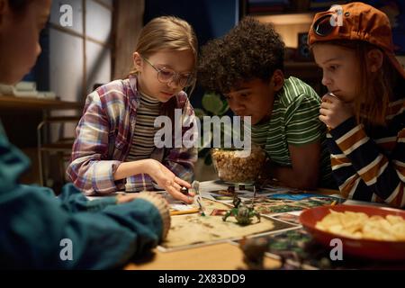 Gruppe ernsthafter interkultureller Kinder, die sich über den Tisch bücken, während einer von ihnen mit Karten und Figuren Würfel auf das Spielbrett wirft Stockfoto