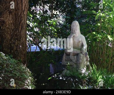 Eine Steinstatue einer sitzenden Figur, umgeben von üppigem Grün und Bäumen in einer ruhigen Umgebung im Freien. Stockfoto