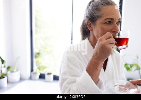 Im Badezimmer zu Hause, reife Kaukasierin trinkt Tee am Fenster Stockfoto