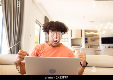 Zu Hause ein birassischer junger Mann mit orangefarbenem T-Shirt, der auf einem Sofa mit einem Laptop anfeuert Stockfoto