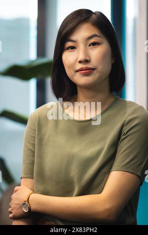 In einem modernen Geschäftsbüro steht die junge asiatische Frau mit überkreuzten Armen und sieht nachdenklich aus Stockfoto