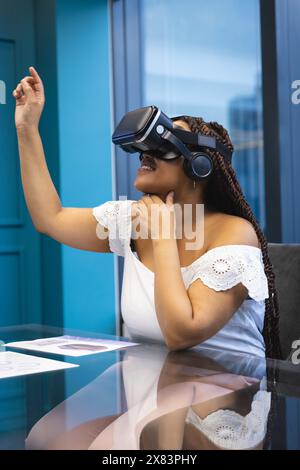 In modernen Büros verwendet junge, birassische Frau mit Zöpfen und schulterfreiem Oberteil ein VR-Headset Stockfoto