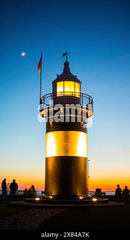 Der schwarz-weiße Leuchtturm, genannt „kleine Preusse“, steht abends vor einem klaren blauen Himmel, einem hellen Leuchtfeuer und aufgehenden Mond Stockfoto