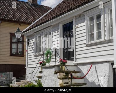 Ein Haus mit schwarzen Türen und Blumen im Vordergrund unter klarem Himmel, neben einer antiken Straßenlaterne, weiße Holzhäuser in grüner Umgebung Stockfoto