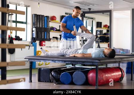 Im Rehabilitationszentrum, birassischer männlicher Physiotherapeut und Patient, der bei Beinübungen assistiert Stockfoto