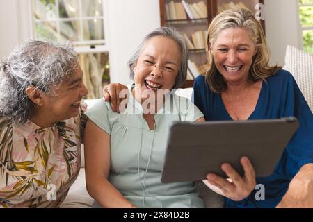 Verschiedene ältere Freundinnen, die zu Hause lachen, eine hält ein Tablet Stockfoto