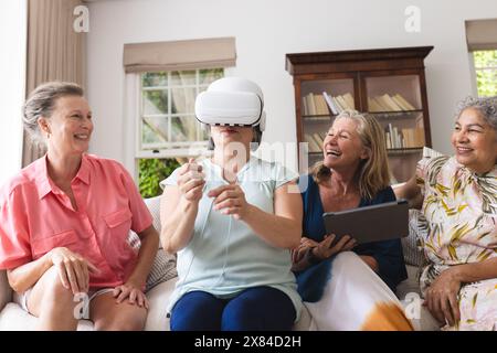 Verschiedene ältere Freundinnen, die zu Hause ein VR-Headset ausprobieren und gemeinsam lachen Stockfoto