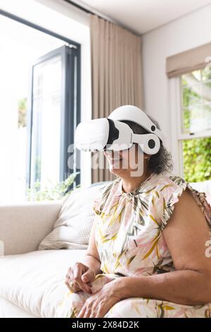 Ältere birassische Frau, die zu Hause ein Virtual-Reality-Headset trägt und auf der Couch sitzt Stockfoto