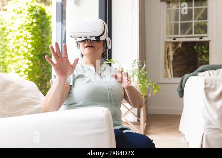 Eine asiatische Seniorin, die zu Hause ein Virtual-Reality-Headset trägt und auf einer Couch sitzt Stockfoto
