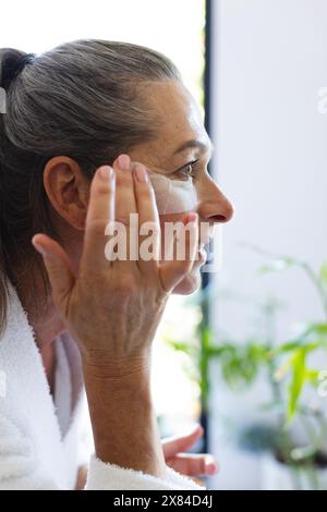 Im Badezimmer zu Hause, reife kaukasische Frau in weißem Gewand, die Creme auf ihr Gesicht aufträgt Stockfoto