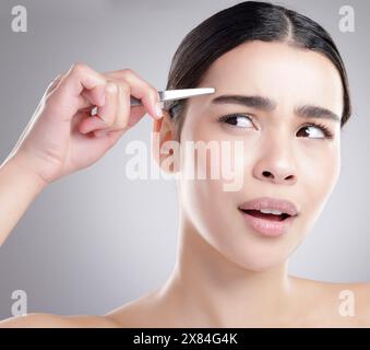 Frau, Schmerz und Augenbrauenpinzette im Studio mit Hautpflege-, Kosmetik- und dermatologischen Anliegen. Junges Modell oder Person mit Zupfen für DIY-Haarentfernung Stockfoto