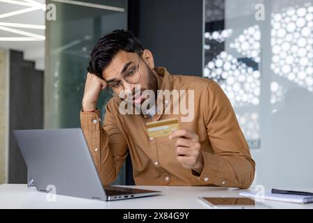Ein Mann in einem braunen Hemd, der an einem Schreibtisch sitzt und gestresst aussieht, während er eine Kreditkarte hält und im Büro einen Laptop benutzt. Stockfoto
