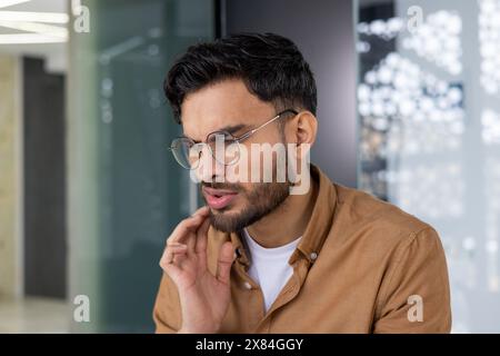 Ein junger Mann mit einer Brille erlebt Zahnschmerzen, während er in einer modernen Büroumgebung sitzt. Stockfoto