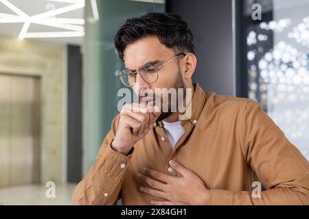 Ein junger Mann hustet mit Hand auf Brust in einer modernen Büroumgebung, was auf Krankheit oder Unwohlsein hinweist. Stockfoto