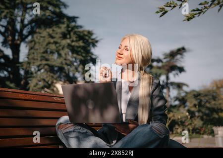 Eine blonde Frau sitzt auf einer Bank mit einem Laptop vor sich. Sie trägt eine graue Jacke und Jeans. Die Szene deutet auf eine lässige und entspannte Atmosphäre hin Stockfoto