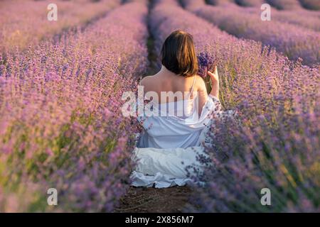 Eine Frau sitzt auf einem Feld mit Lavendelblumen. Sie hält einen Blumenstrauß in der Hand. Die Szene ist ruhig und friedlich, mit der Frau Stockfoto
