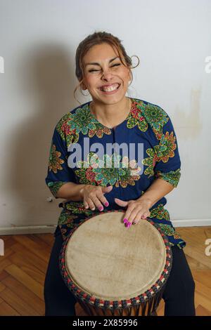 Eine Erwachsene, sehr glückliche Lateinfrau, die zu Hause mit ihrer Djembe-Trommel sitzt. Sie spielt, lächelt und genießt sich selbst. Stockfoto