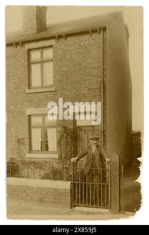 Ursprüngliche Postkarte aus der Zeit des 1. Weltkriegs mit einer typischen flachen Mütze und einem Anzug, stehend vor der Haustür seines typisch britischen roten Ziegelsteinterrassenhauses Nr. 53, typisch britisches Gehäuse aus den 1900er Jahren, durch sein Gartentor, eine Frau aus dem Fenster zwischen den Vorhängen, um 1917, Großbritannien Stockfoto