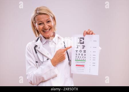 Porträt einer reiferen Ärztin, die Augenuntersuchungen auf grauem Hintergrund durchführt. Stockfoto