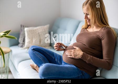 Schwangere Frau sitzt zu Hause auf dem Sofa und sieht sich das Ultraschallbild ihres Babys an Stockfoto