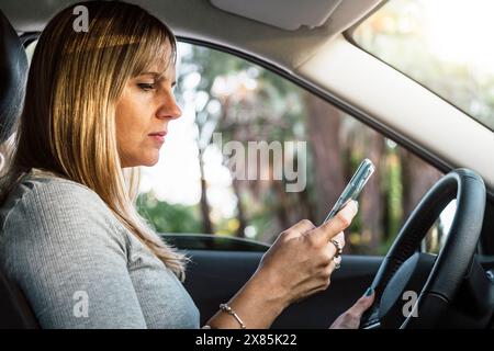 Fahrerin im Auto, die ihr Handy benutzt. Ernsthafte Frau mittleren Alters, die mit einem Smartphone in der Hand in einem Fahrzeug sitzt. Stockfoto