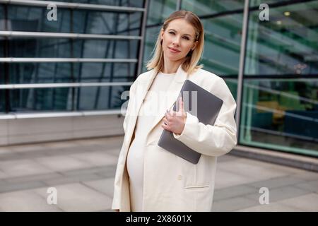 Schöne schwangere blonde Frau in einem weißen Business-Anzug mit Blazer steht draußen mit einem Laptop in der Nähe eines Firmengebäudes und zeigt Professionalität Stockfoto