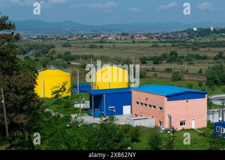 Eine ländliche Industrieanlage mit leuchtend gelben Tanks, blauen Strukturen und sanften grünen Hügeln mit einem entfernten Dorf. Stockfoto