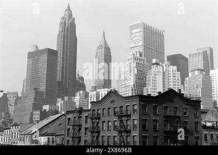 Blick auf Manhattan, während sich im Hintergrund das Empire State Building und 40 Wall Street erheben, liegt im Vordergrund ein Wohngebäude aus Ziegelsteinen mit den typischen eisernen Feuerleitern an der Außenfassade, New York 1967. Blick auf Manhattan, mit dem Empire State Building und der Wall Street 40 im Hintergrund und einem Backsteingebäude mit dem typischen Eisenfeuer auf der Fassade im Vordergrund, New York 1967. Stockfoto