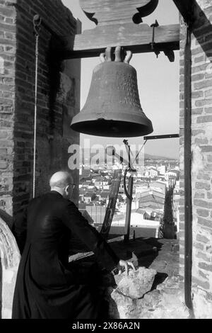 Ein Geistlicher blickt an der Glocke eines Glockenturms vorbei auf die umliegende Ortschaft von Calella an der Costa del Maresme, Katalonien 1957.Ein Geistlicher blickt an der Glocke eines Glockenturms vorbei an der umliegenden Ortschaft Calella an der Costa del Maresme, Katalonien 1957. Stockfoto