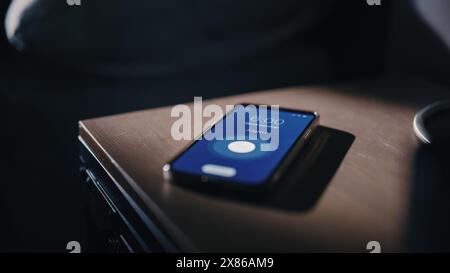 Smartphone-Wecker-App-Ringe zum Aufwachen, Bildschirm zeigt „Guten Morgen“ und die Uhrzeit von sechs Uhr am Morgen an. Nahaufnahme: Handyklingeln. Apartment mit Nachttisch und Schlafzimmer Stockfoto