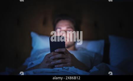Der kaukasische Mann benutzt nachts ein Smartphone im Bett. Gutaussehender Kerl, der in sozialen Medien surft, Nachrichten liest, Online-Shopping macht, spät in der Nacht mit Freunden chatten kann. Konzentrieren Sie sich auf das Handhalten des Mobiltelefons Stockfoto