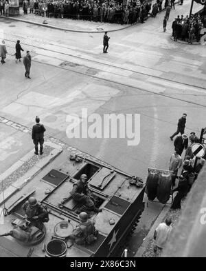 Ein Geschäftsmann zieht einen voll-behängten Geraderobenständer vor einem Panzer der US-Army auf den Bürgersteig. Im Hintergrund haben sich interessierte Passantinnen und Passanten zahlreich auf den Bürgersteigen versammelt; einige überqueren die Straße, Berlin 1961. - Originale Bildbeschreibung: "Die Damenoberbekleidung arbeitet unter den Augen von amerikanischen Soldaten. Trotz der gespannten Situation geht die Arbeit der West-Berliner wie immer voran.- Übergriffe werden abgewiesen - zu einem schweren Grenzzwischenfall kam es auch heute wieder an der Friedrich-/Ecke Kochstraße, als zwei ame Stockfoto