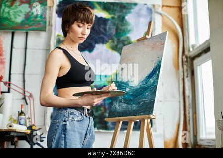 Eine Frau in einem schwarzen Tanktop konzentriert sich auf das Malen auf einer Staffelei. Stockfoto