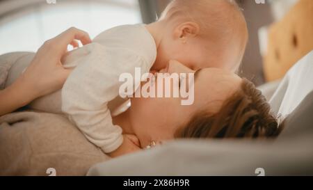 Liebende junge, liebevolle Mutter, die zu Hause ein entzückendes Baby spielt und pflegt. Lächelnde schöne Mutter und niedliches kleines Kleinkind kuscheln im Schlafzimmer. Mütterliche Zärtlichkeiten. Stockfoto