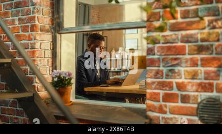 Porträt eines jungen gutaussehenden Schwarzen Mannes, der an einem Schreibtisch sitzt und an einem Laptop arbeitet. Freiberuflicher Journalist, der einen Artikel aus der Ferne aus dem Home Office schreibt. Stilvolles Apartment aus dem Fenster. Stockfoto