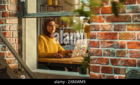 Junge muslimische Studentin mit Laptop zu Hause. Teilnahme an Fernschulungen während des Tages. Arbeiten am Computer, Schreiben eines Aufsatzes. Bogenaufnahme von außerhalb des Fensters. Stockfoto