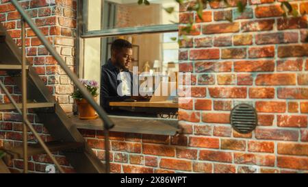 Junger Schwarzer, der zu Hause ein Notebook benutzt. Inspirierende Fernarbeit während des Tages. Spezialist, der am Computer arbeitet, einen kreativen Projektplan schreibt. Von außerhalb des Fensters geschossen. Stockfoto