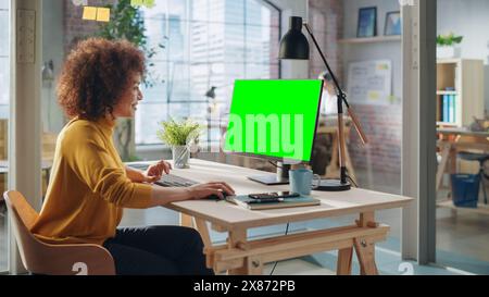 Porträt eines aufgeregten multiethnischen Spezialisten, der in Creative Agency arbeitet. Stilvolle ausdrucksstarke Frau im gelben Jumper mit Desktop-Computer mit grünem Bildschirm Mock Up Display. Stockfoto